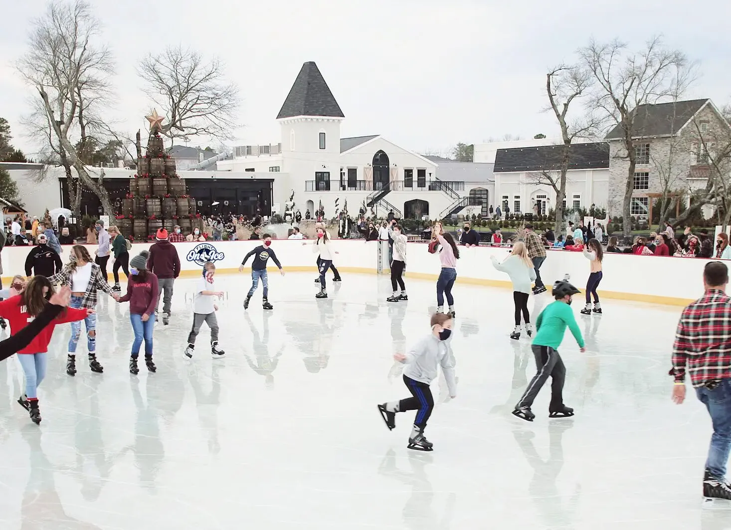 Skaters filling the ice rink at Renault Winery, with the white winery building and the barrel Christmas tree behind the boards. — Flight on Ice at Renault Winery