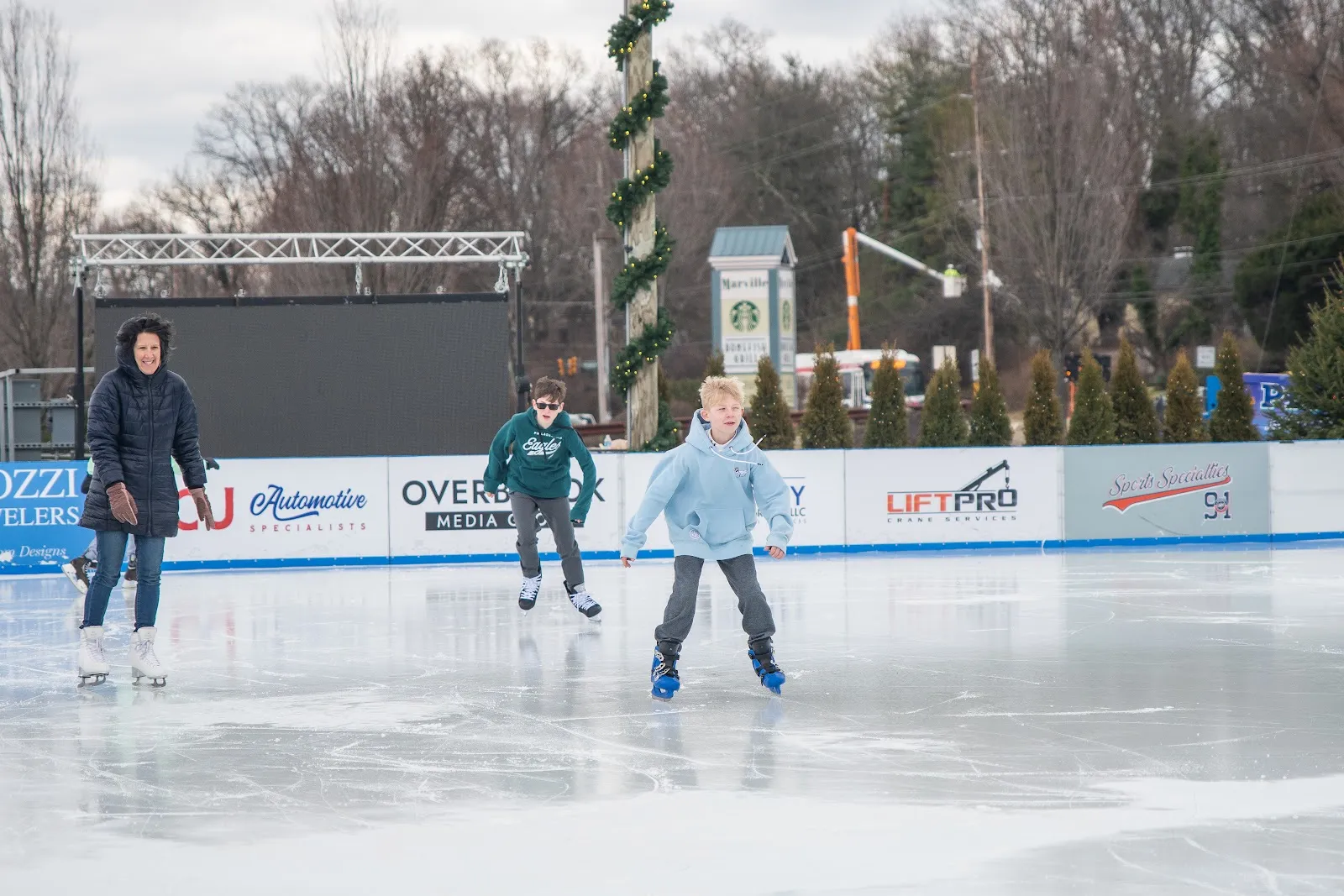Skaters crossing the boarded outdoor rink at Newtown Square on a cold afternoon, with sponsor boards along the far side. — Flight on Ice Newtown Square