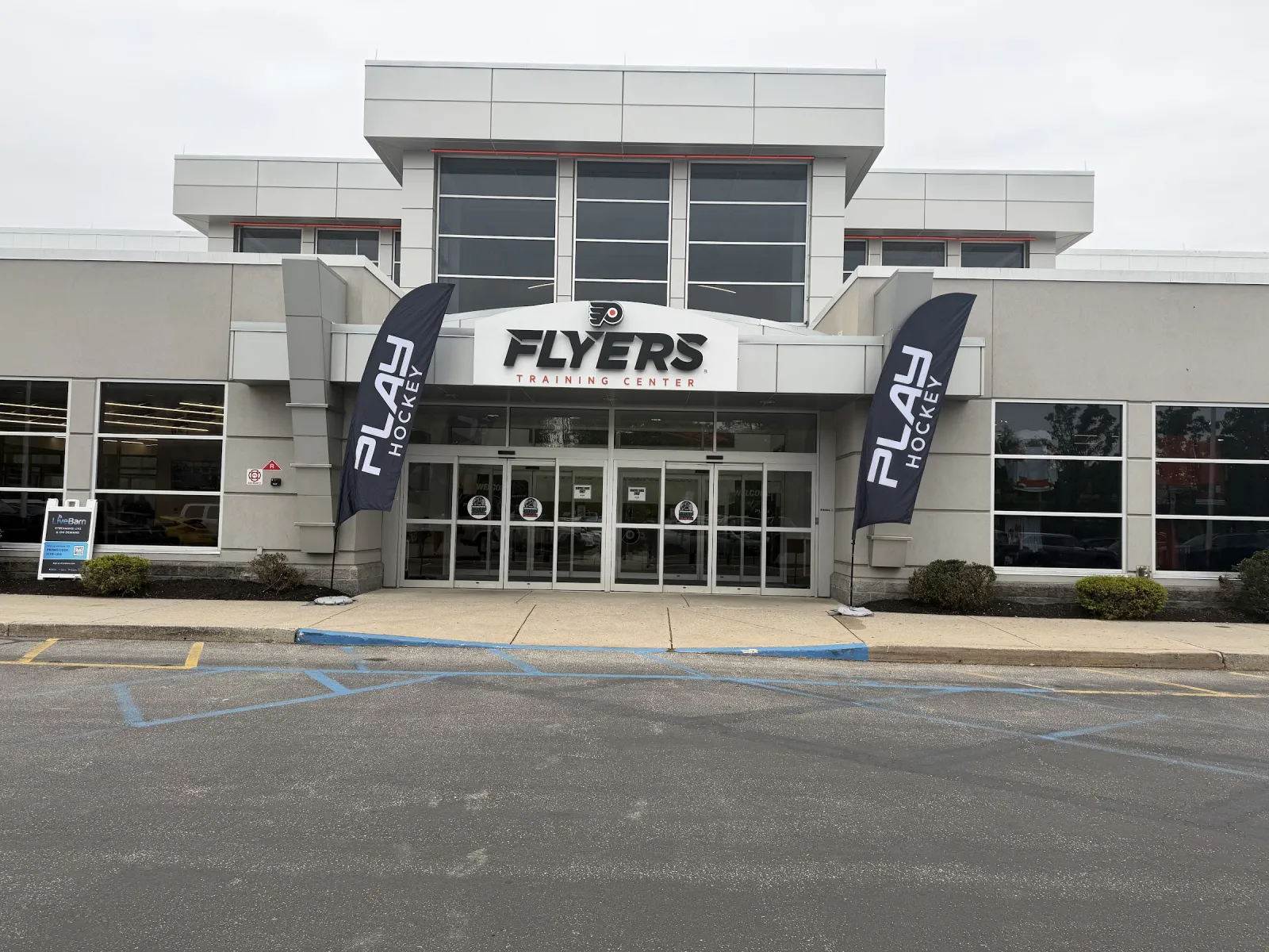 The grey two-storey entrance with a Flyers sign over the glass doors and Play Hockey banners on either side, seen from the parking lot. — Flyers Training Center