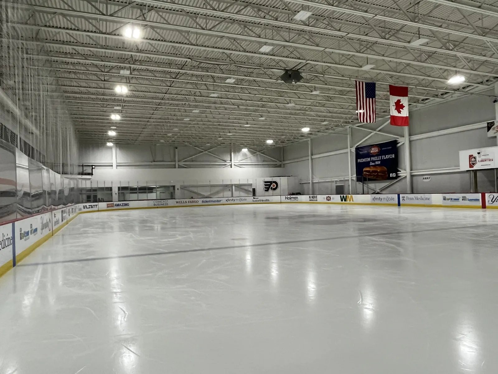 An empty ice sheet under a white steel roof, with American and Canadian flags and a scoreboard at the far end and sponsor boards along the dasher. — Flyers Training Center
