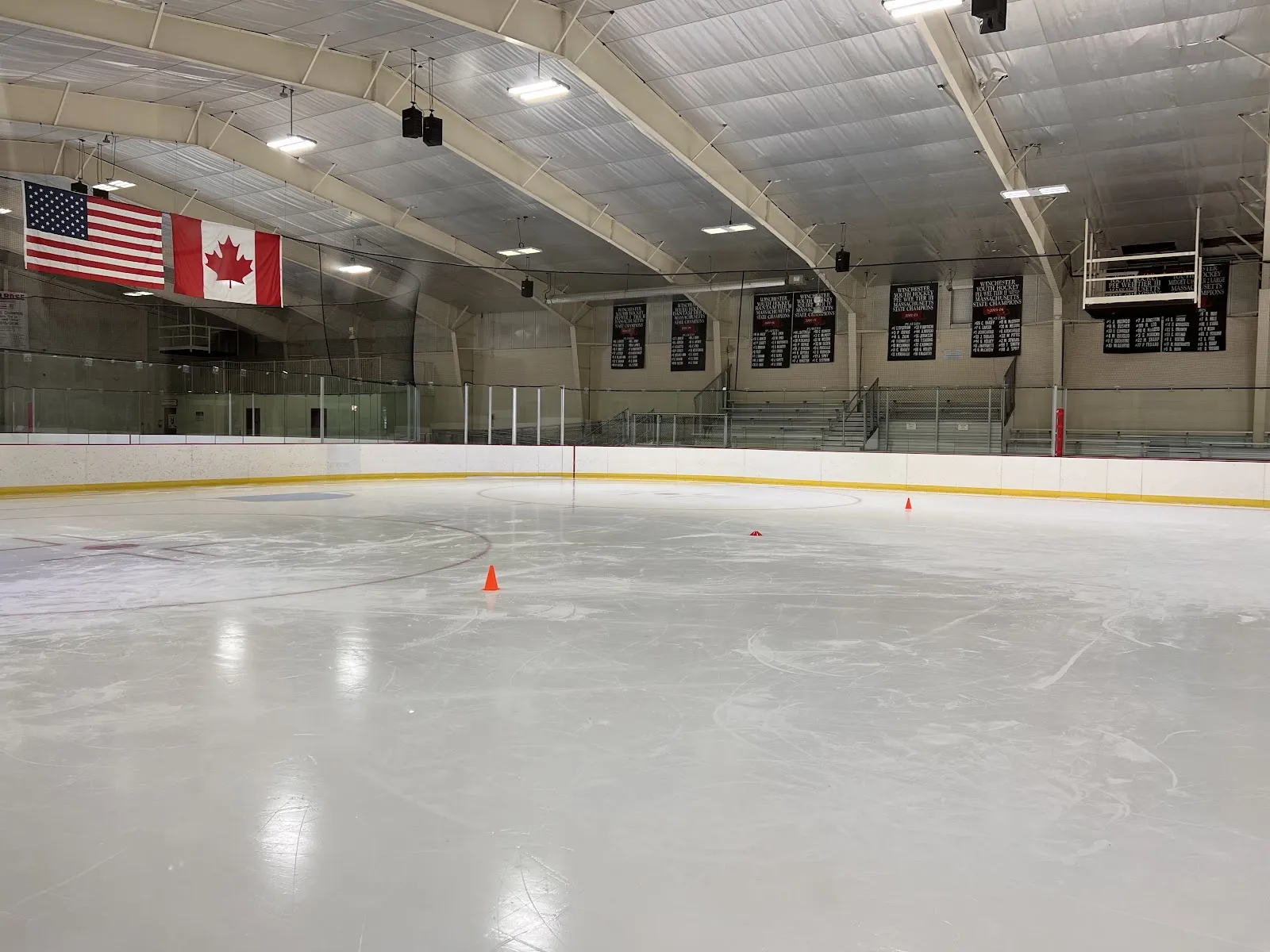 The empty Flynn Rink ice under its arched roof, with U.S. and Canadian flags and team banners at the far end. — Flynn Memorial Rink