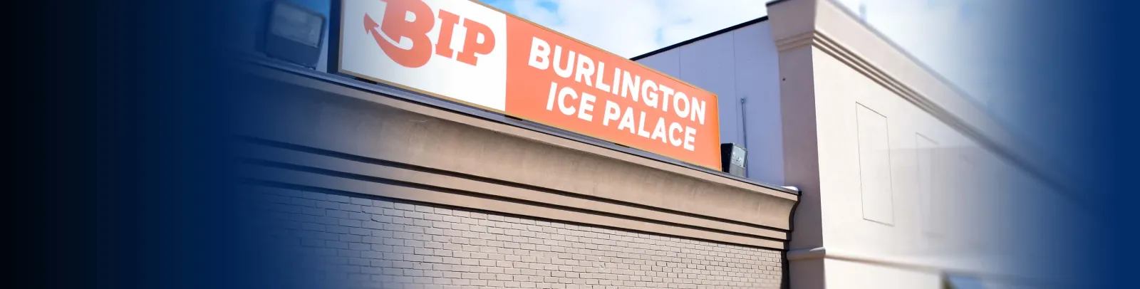 Upper-wall sign reading "BIP BURLINGTON ICE PALACE" (orange and white) mounted above a tan cornice and brick facade, corner of the building visible against a blue sky. — Burlington Ice Palace