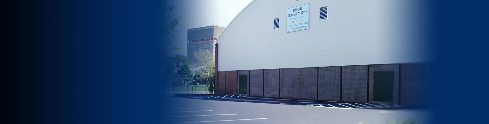 Exterior de un edificio estilo nave Quonset con un letrero en la pared que dice "SIMONI MEMORIAL RINK, Operated by FMC Arena 4", muro inferior de ladrillo, estacionamiento y un terreno cercado al frente, y el perfil de Boston visible a lo lejos. — Simoni Skating Rink