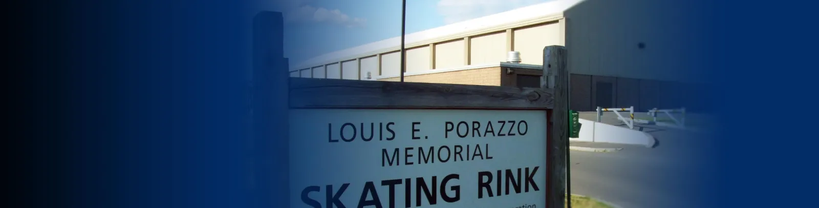 Wooden entrance sign reading "LOUIS E. PORRAZZO MEMORIAL SKATING RINK" in front of the building's tan-and-brick exterior wall, a swing gate and paved lot visible behind. — Porrazzo Skating Rink
