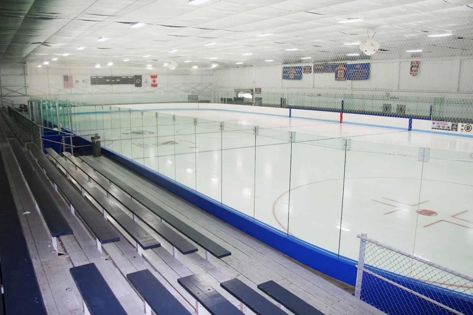 Empty ice sheet viewed from the stands, glass boards, blue kick plates, penalty boxes and team banners on the far wall, no people on the ice. — Porrazzo Skating Rink