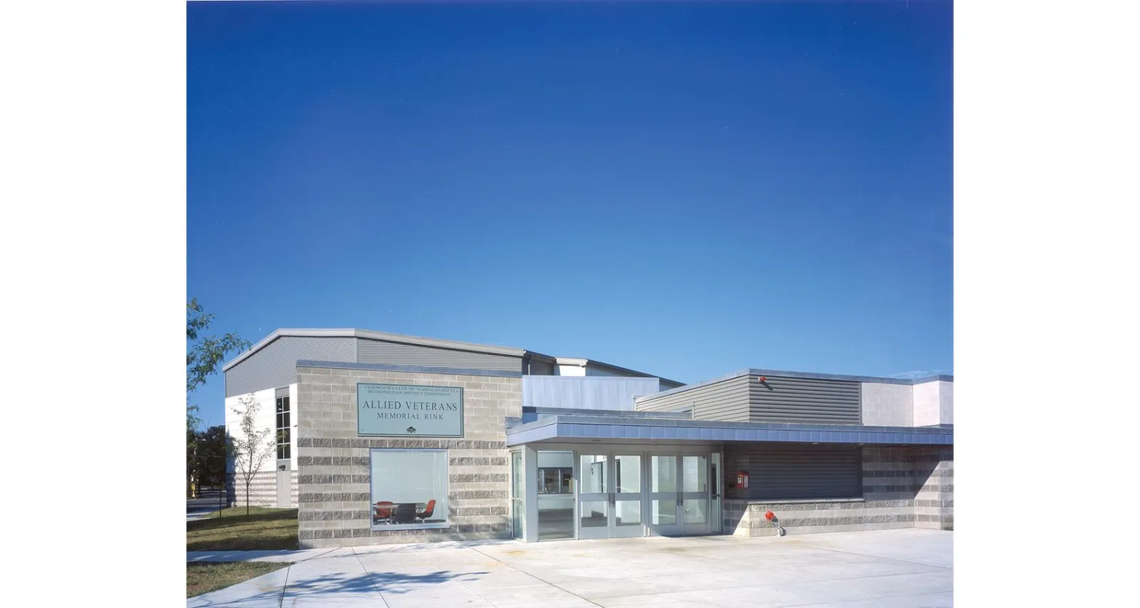 Building exterior with a mounted sign reading "Allied Veterans Memorial Rink" (Commonwealth of Massachusetts, Metropolitan District Commission) beside the glass entrance, blue sky. — Allied Veterans Memorial Rink