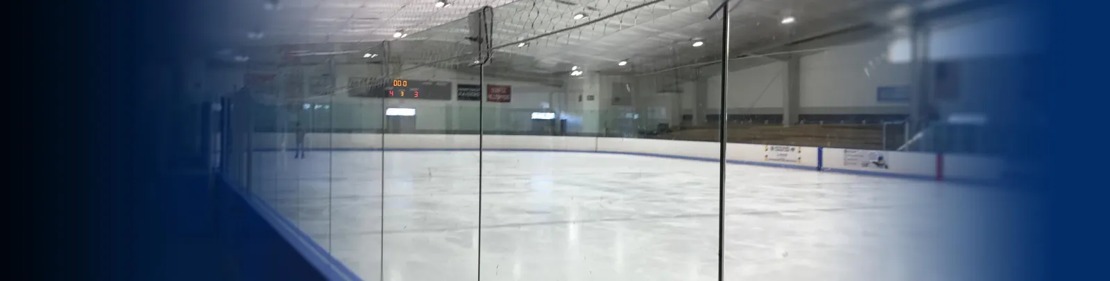 Empty ice sheet seen through the glass along the boards, with netting overhead, a scoreboard on the far wall and bleachers on the right. — Driscoll Skating Arena