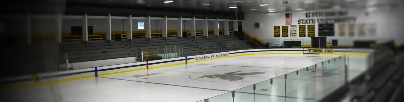 Empty indoor ice sheet with a black-and-gold eagle logo at center ice, a rink-board sign reading "Captain Kenneth W. Gaetz, Sr. Arena" and "Fitchburg State", bleacher seating and championship banners on the wall behind. — Wallace Civic Center