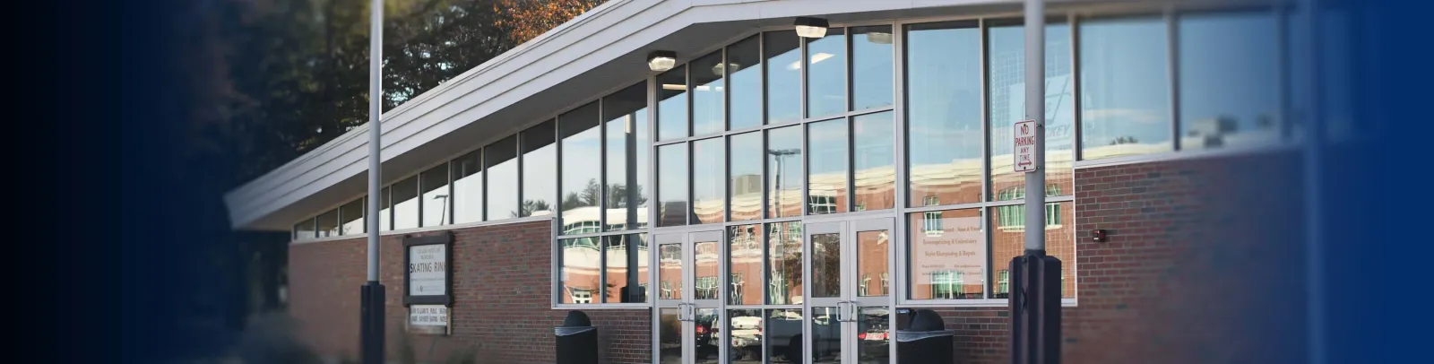 Front entrance of a brick building with a sloped white roofline, a sign by the door reading "Collins-Moylan Memorial Skating Rink", and glass doors under a glass-walled lobby. — Collins/Moylan Arena