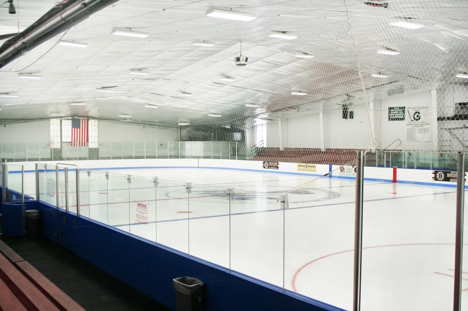 The empty Collins-Moylan Arena rink from the bleachers, blue boards and glass, a flag and banners on the far wall under a white roof — Collins/Moylan Arena