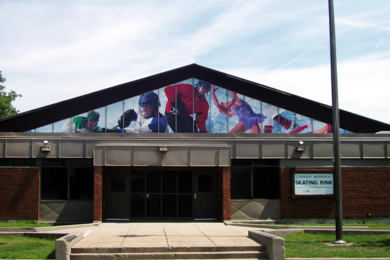 Frente de Connery Memorial Skating Rink, un edificio bajo de ladrillo con un frontón puntiagudo que tiene gráficos deportivos en las ventanas sobre la entrada y un letrero que dice "CONNERY MEMORIAL SKATING RINK / Operated by Facility Management Corporation". — Connery Skating Rink