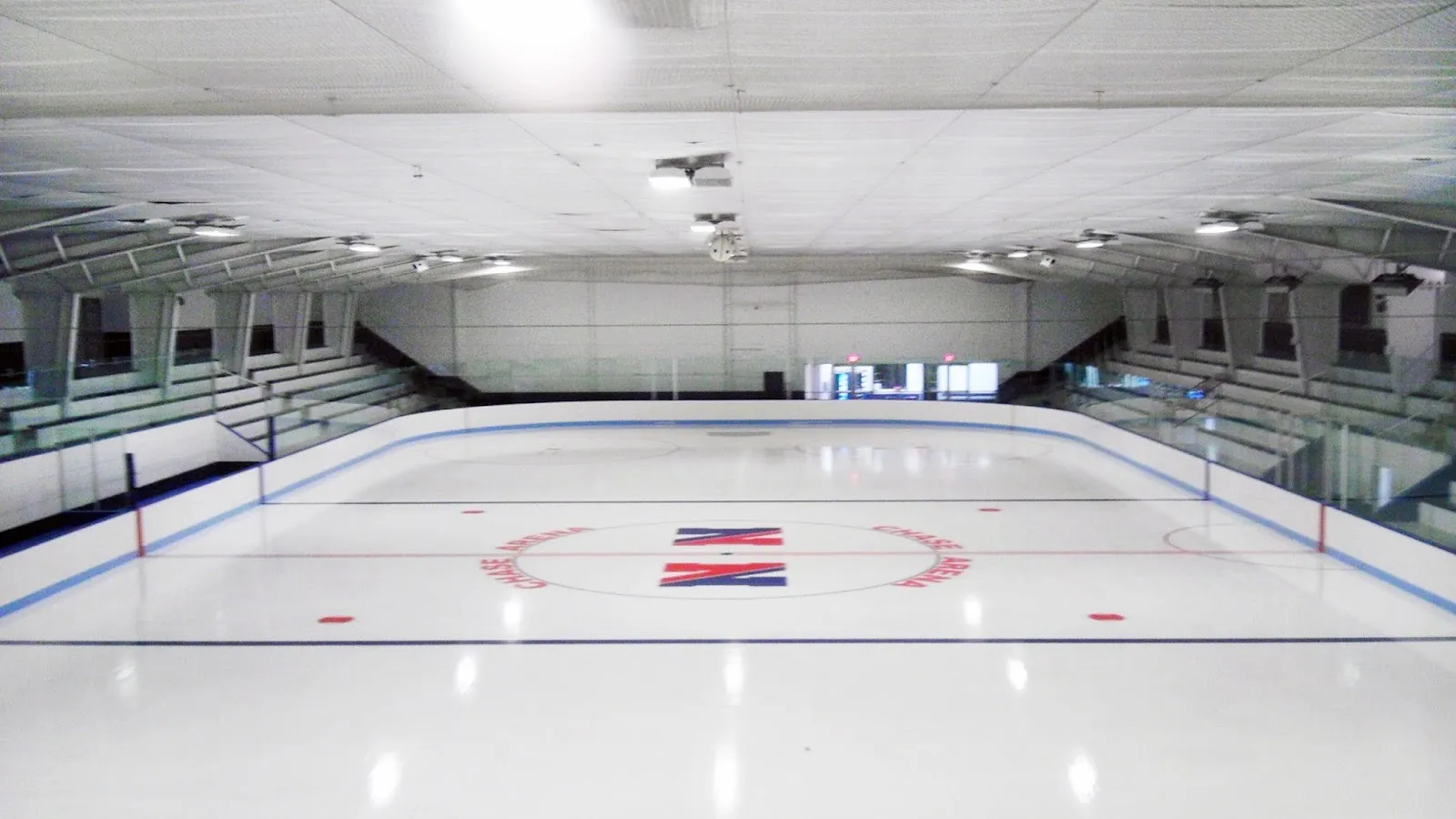 La pista vacía de William L. Chase Arena desde las gradas, con Chase Arena y la N de Natick marcadas en el centro del hielo entre gradas — William L. Chase Arena