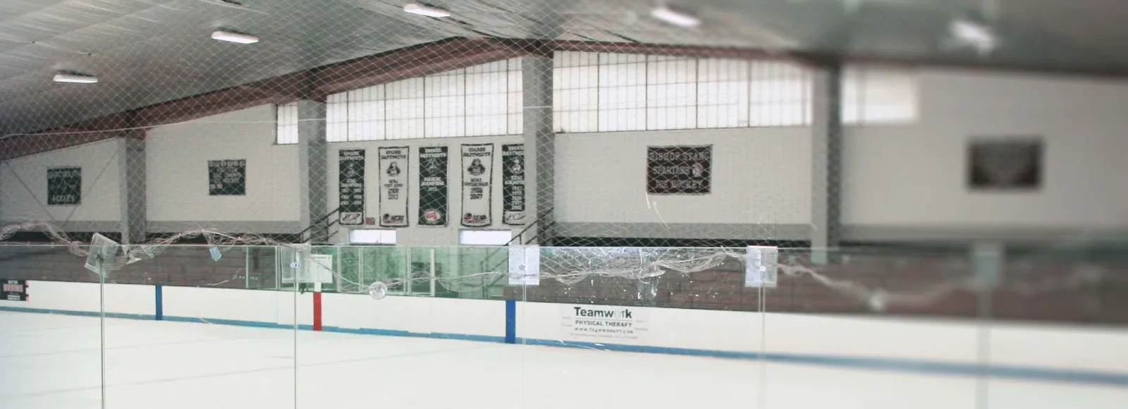 Interior view of the ice sheet from behind the glass and netting, showing part of the boards and the championship banners (Dartmouth Hockey, Bishop Stang Ice Hockey) on the end wall; no people. — Hetland Arena