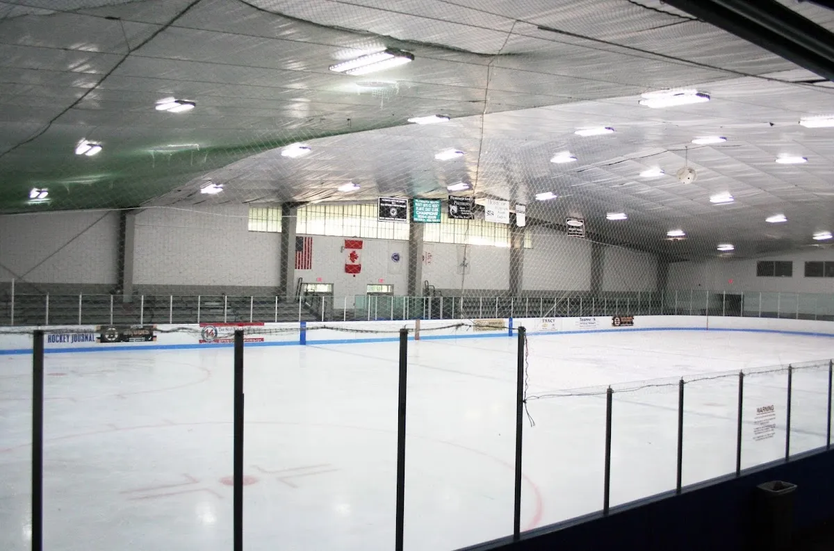 Empty full ice sheet seen from the stands, with the boards, blue center line, hanging scoreboard, an American flag and Canadian flag on the end wall, and the ceiling lighting structure. — Armstrong Arena