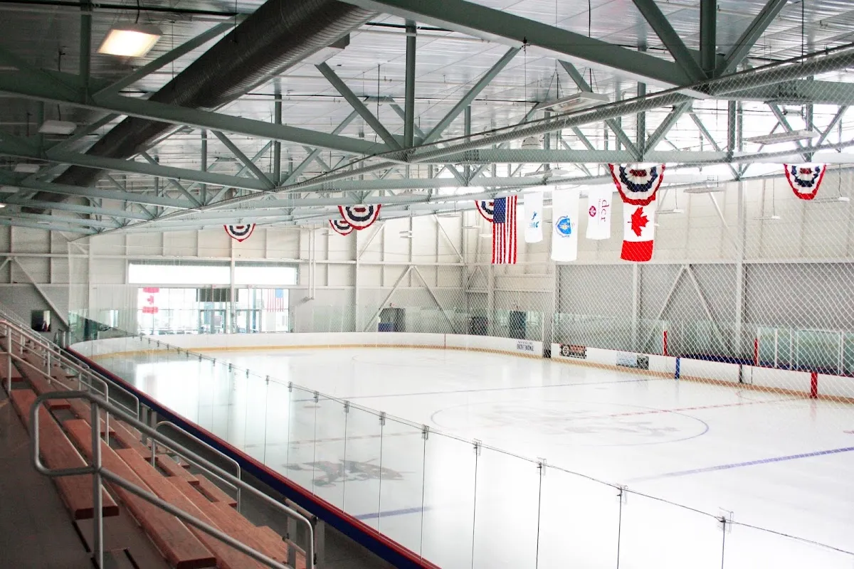 Empty full ice sheet under exposed steel roof trusses, with American, Canadian and organization pennants hanging from the ceiling and bleacher seating in the foreground; a "dcr" (Massachusetts Department of Conservation and Recreation) banner is visible among the hanging flags. — Cronin Skating Rink