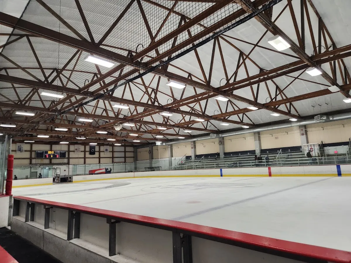 Empty full ice sheet from rinkside, showing the wooden roof trusses, an overhead scoreboard, hanging banners, bleacher seating and a large circular rink logo at center ice. — Veterans Memorial Rink