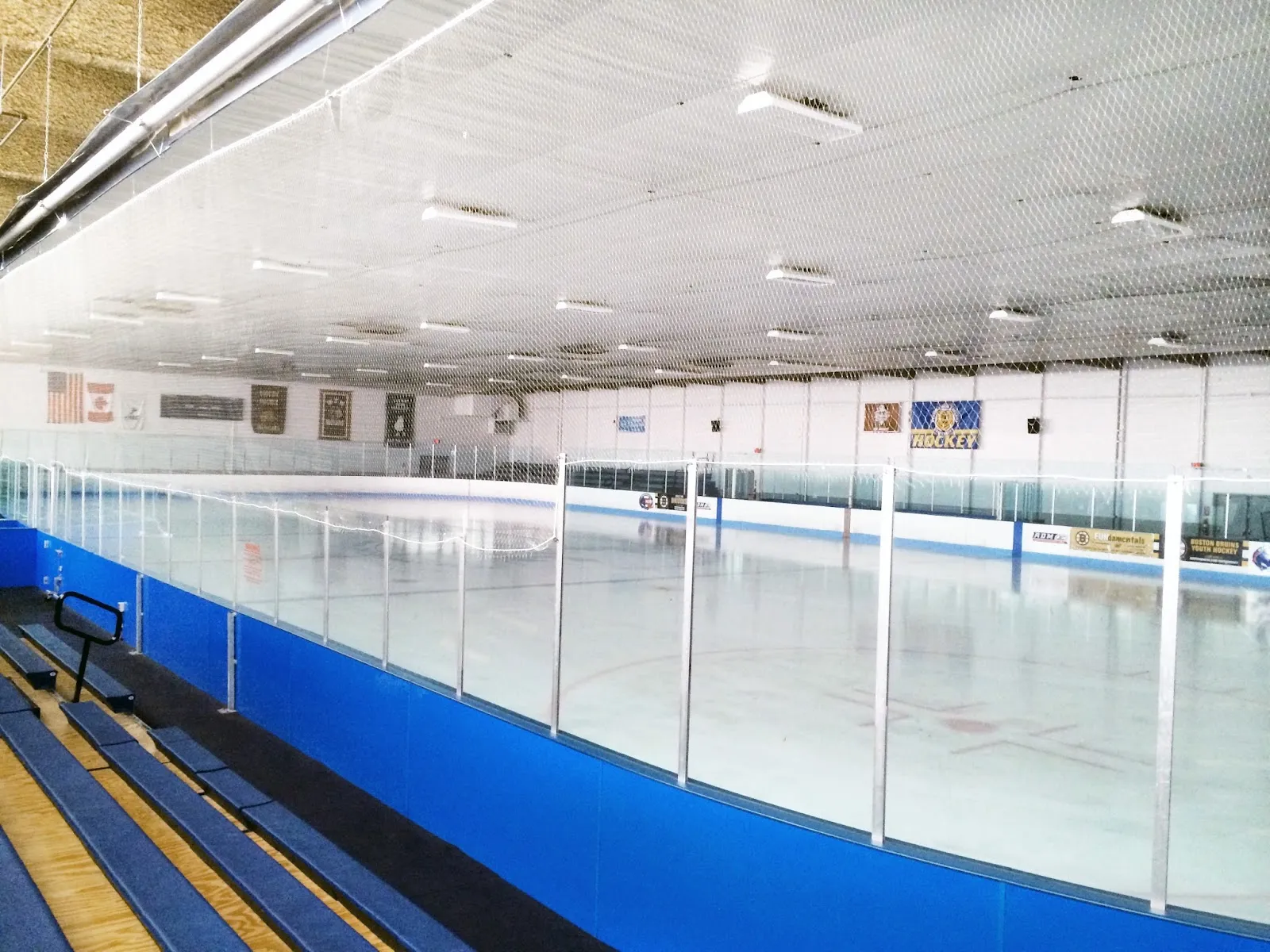 Empty ice sheet with blue dasherboards, wall-mounted youth-hockey and championship banners, and bleacher seating along one side. — Smead Arena