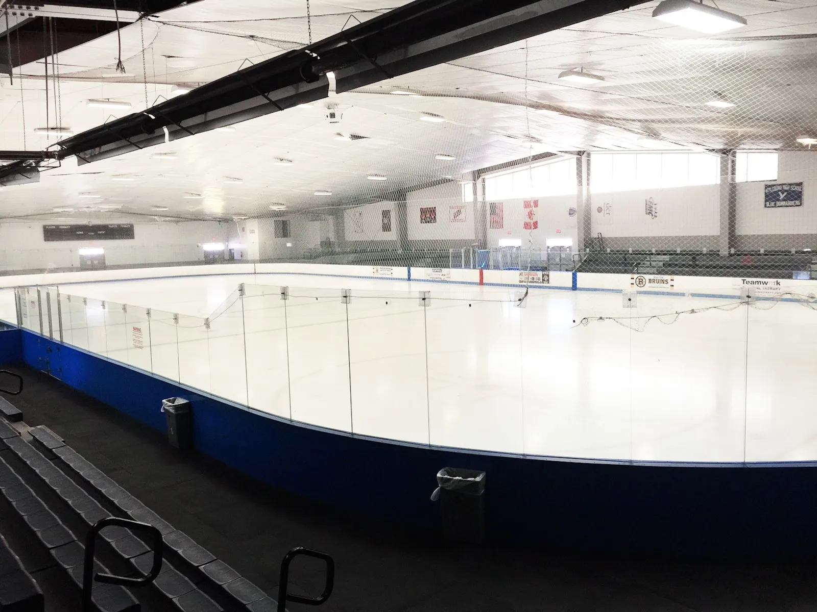 Pista de hielo vacía bajo un techo a dos aguas con un marcador, cristal y vallas, y mantas de equipos en las paredes, incluidas las de Attleboro High School y Boston Bruins Youth Hockey. — Aleixo Arena