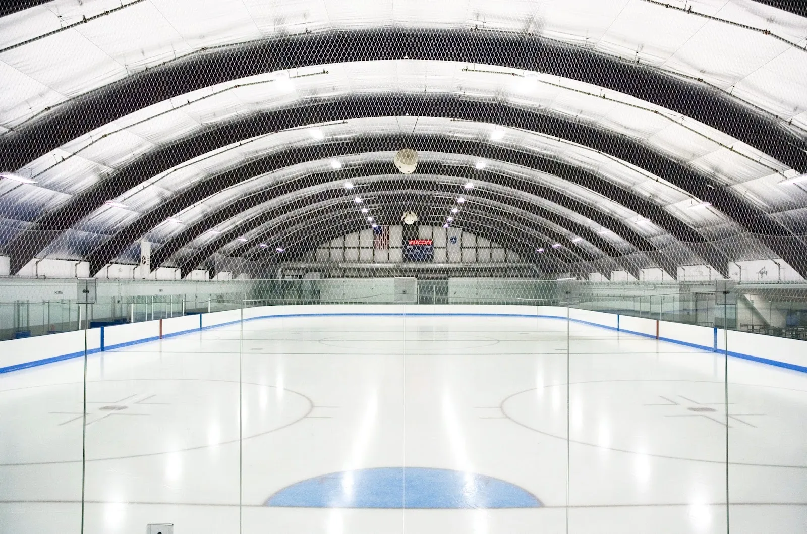 Empty ice sheet inside an arched Quonset-hut-style rink building, with an overhead scoreboard, American flag, and blue dasherboards. — Jim Roche Community Arena