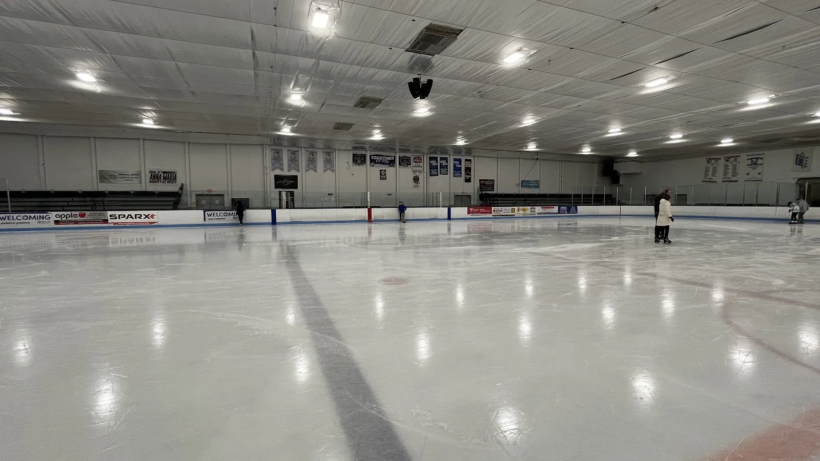 Pista casi vacía durante el patinaje público, con algunos patinadores a lo lejos, anuncios de patrocinadores en las vallas y mantas de equipos en las paredes. — Buffone Skating Arena