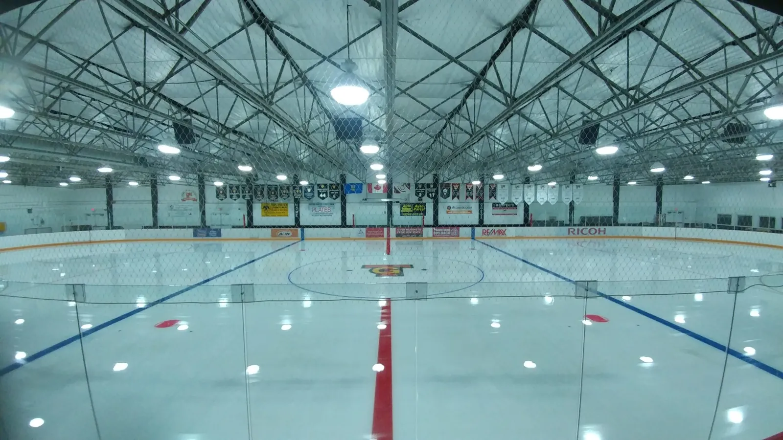 La arena vacía desde detrás del cristal en el centro de la pista: una línea central roja, líneas azules y un logotipo en el centro de la pista bajo una red de armaduras de acero en el techo, con mantas de equipos y una bandera de Canadá en la pared del fondo. — Forbes Innes Arena