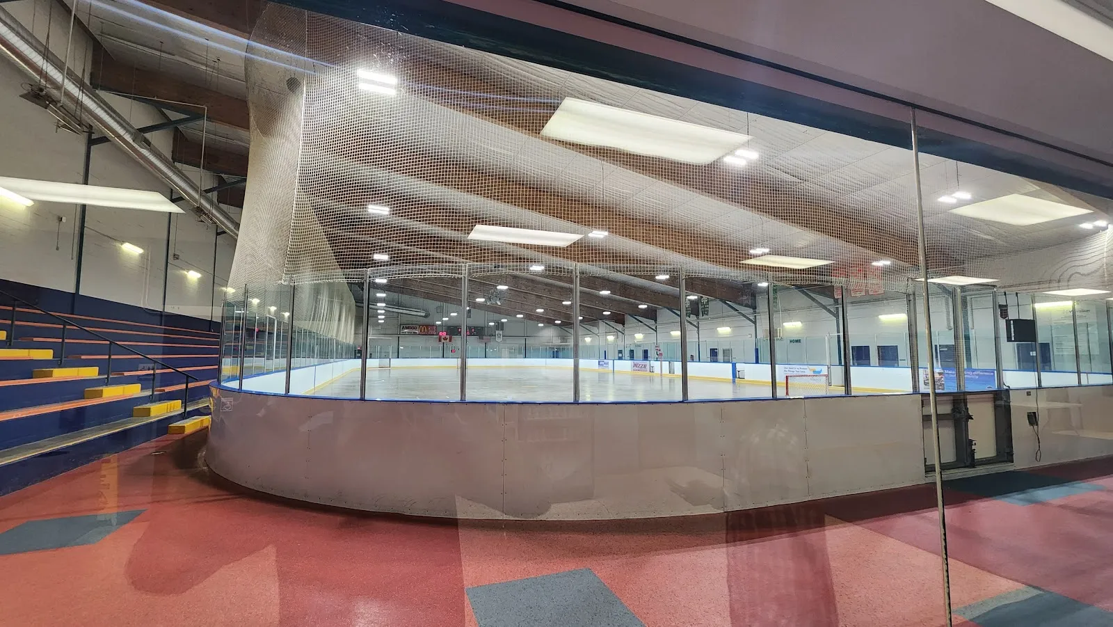 El hielo vacío de Forest Glade Arena visto a través del cristal del vestíbulo, con la red de seguridad, el techo de madera y las gradas al costado — Forest Glade Arena