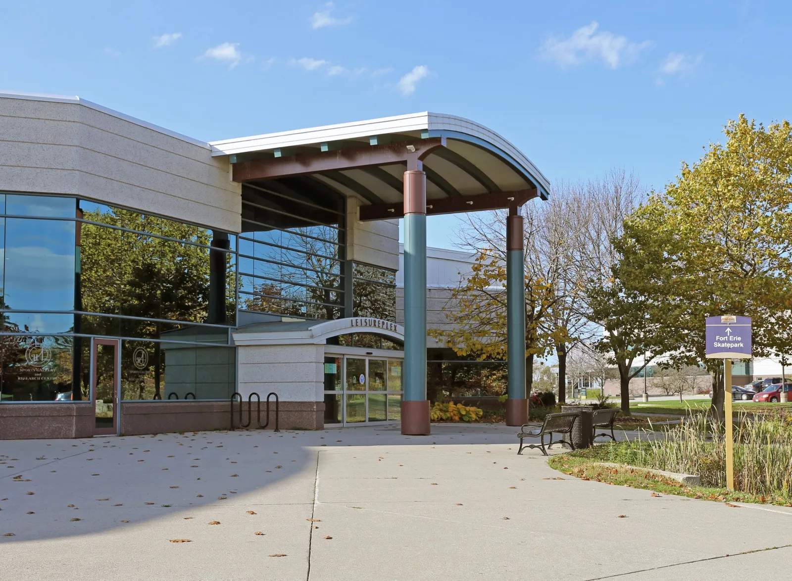 La entrada del Leisureplex con su letrero LEISUREPLEX, un pasillo techado sobre columnas, bancas y árboles otoñales junto a un letrero que señala el Fort Erie Skatepark. — Fort Erie Leisureplex