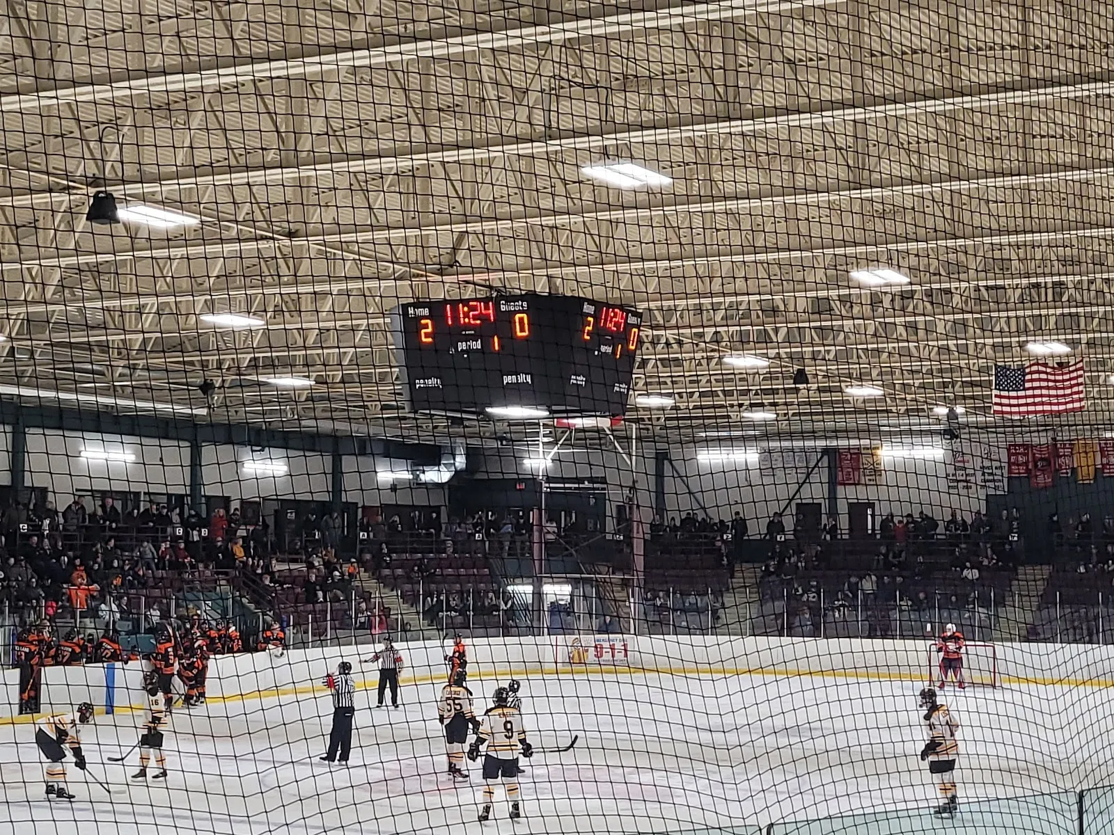 Un partido visto desde un barandal junto a la arena, con jugadores repartidos por la pista y unas largas gradas de asientos color granate alrededor, bajo un techo de armaduras de acero. — Fort Erie Leisureplex
