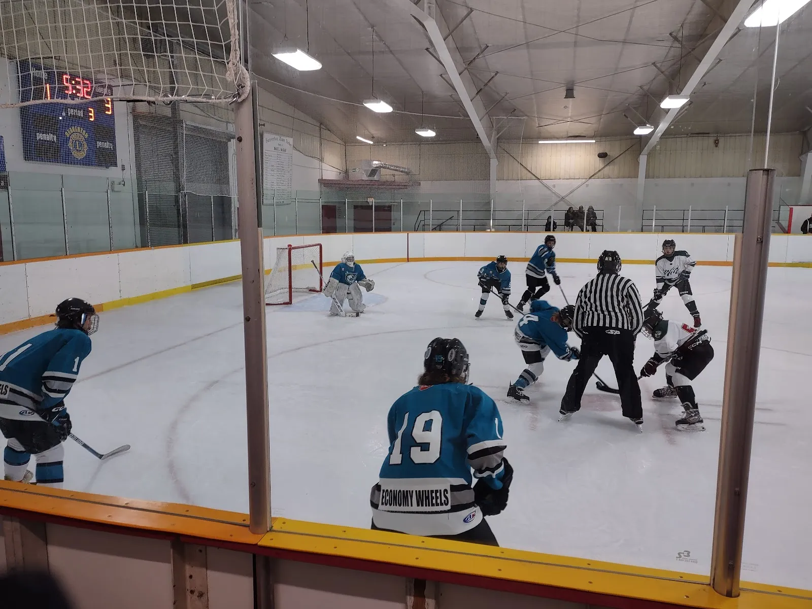 A game on the ice at the Foster Hewitt Memorial Community Centre, with a face-off at the near circle, the scoreboard and netting behind the boards. — Foster Hewitt Memorial Community Centre