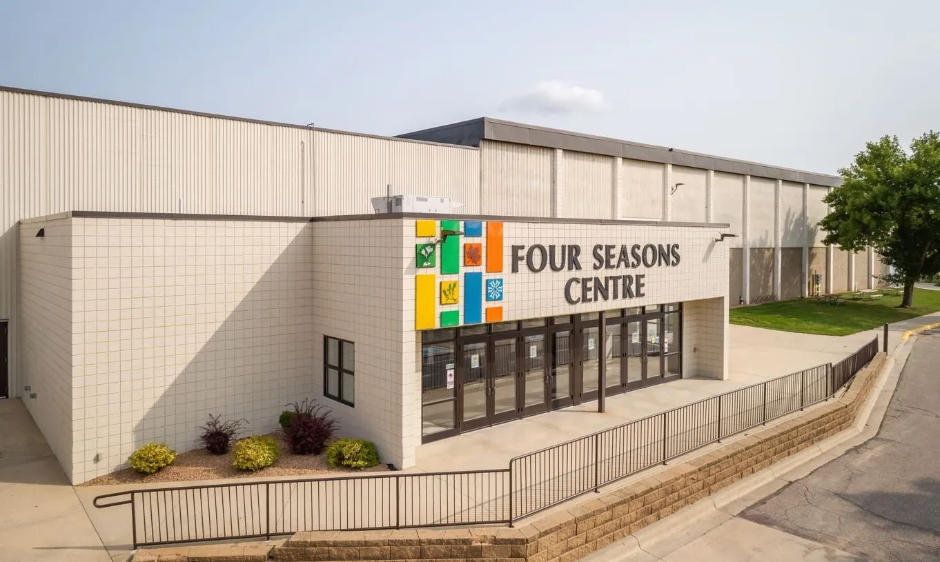 La entrada principal del Four Seasons Centre: un edificio de bloque color crema con las letras 'FOUR SEASONS CENTRE' y paneles de colores sobre las puertas de vidrio, con una rampa y un barandal al frente. — Steele County Four Seasons Centre