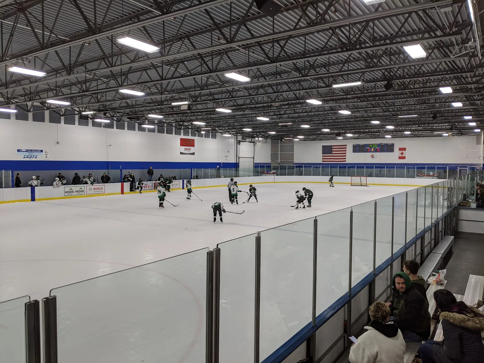 Una pista completa durante un partido de hockey juvenil, vista a lo largo del vidrio desde el extremo de las gradas, bajo un techo luminoso de armaduras de acero con banderas en la pared del fondo. — Steele County Four Seasons Centre