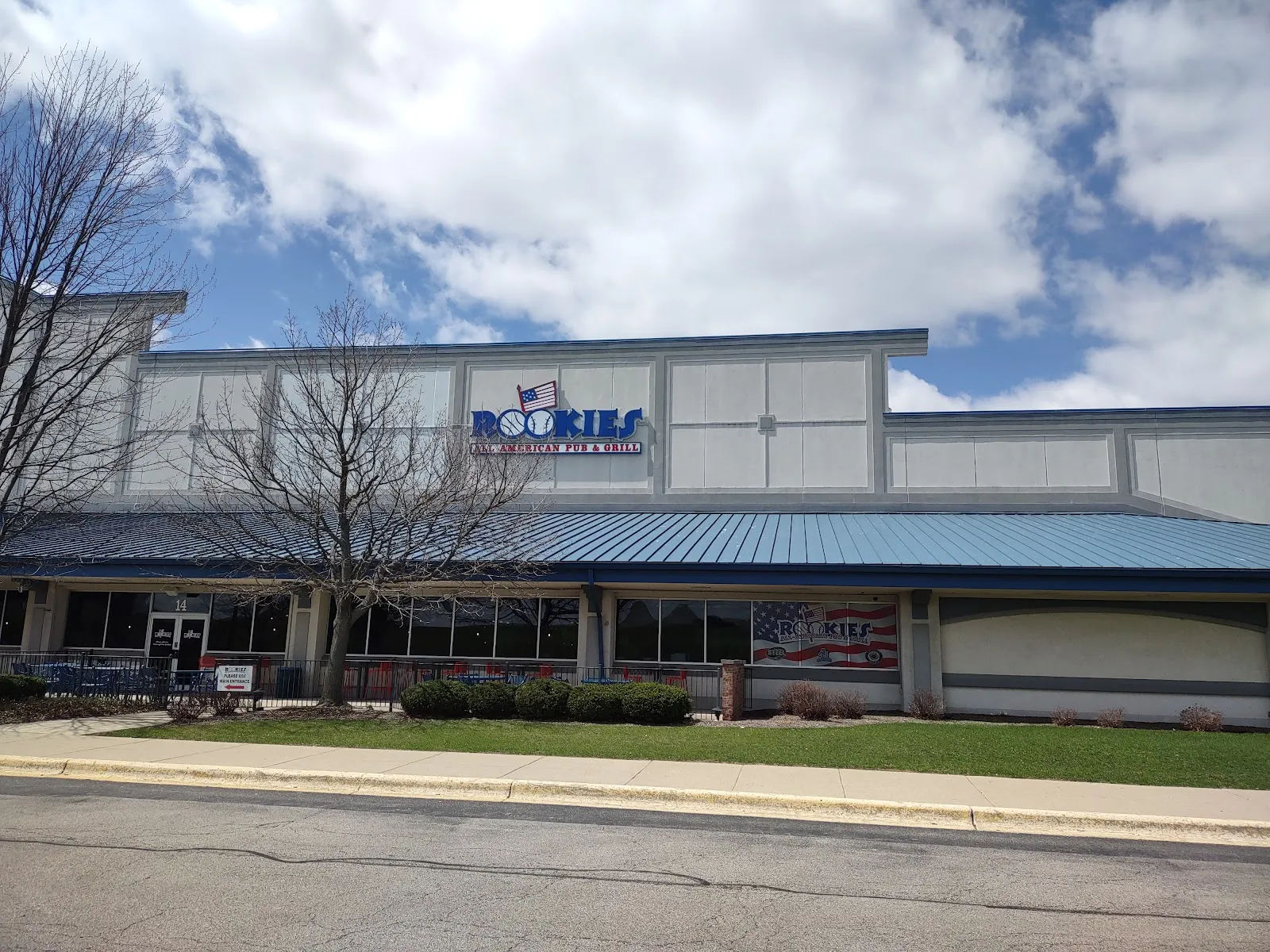 The arena's long pale front under a blue roof strip with a Rookies sign above the entrance, seen across the lawn under a cloudy blue sky. — Fox Valley Ice Arena
