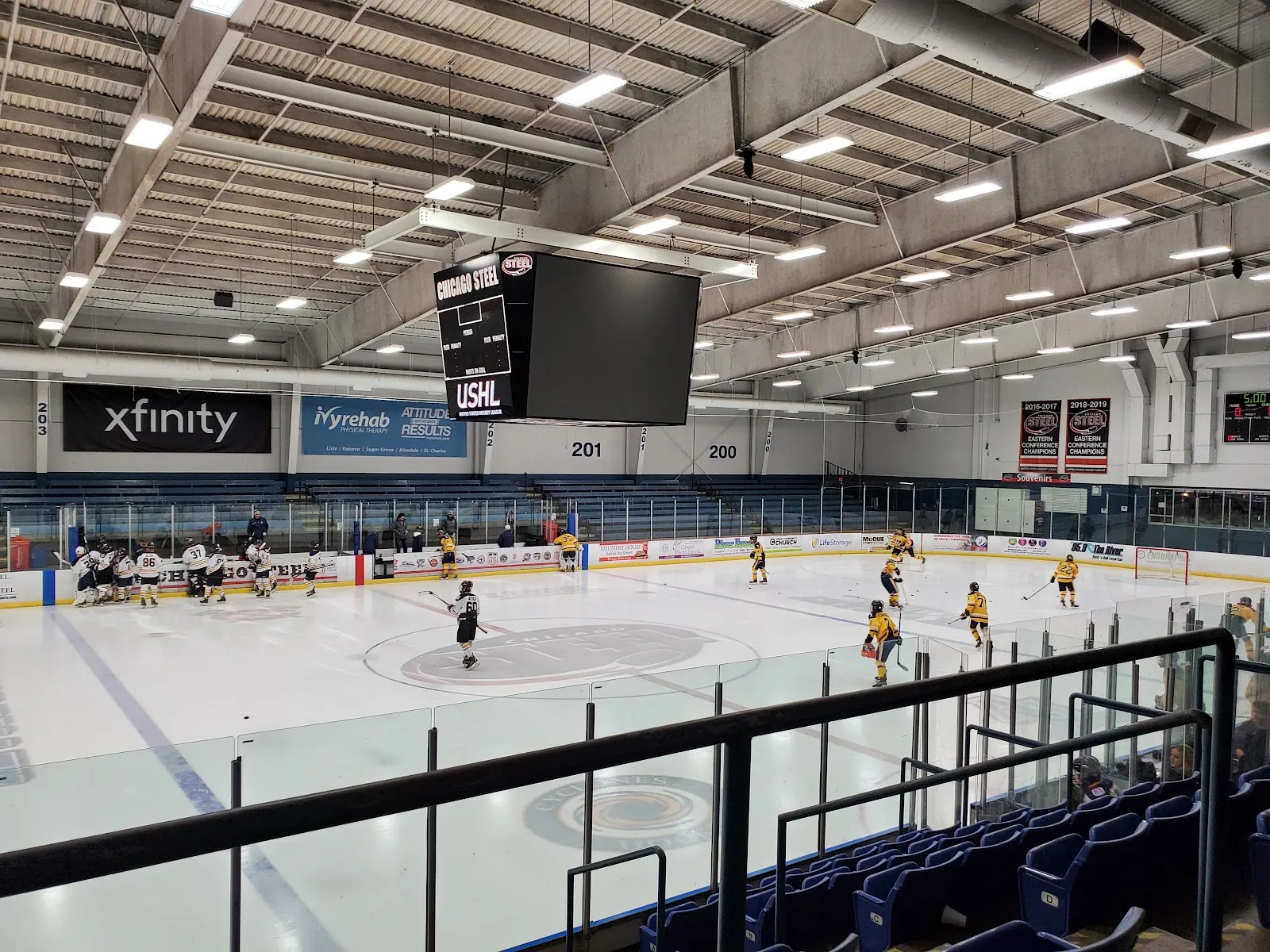 A pre-game view of the sheet from the stands, players spread across the ice under a long ceiling of lights with scoreboards and banners along the far boards. — Fox Valley Ice Arena