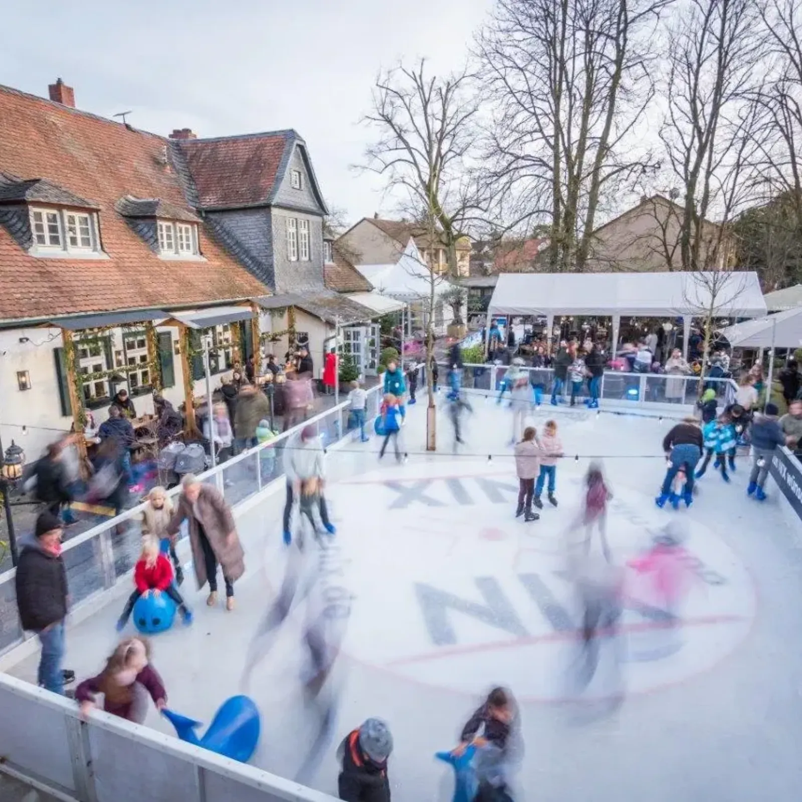 Skaters and children on the open-air ice rink at the Frankfurter Haus in daylight, with the half-timbered buildings and market tents around the rink. — Eislaufbahn im Frankfurter Haus