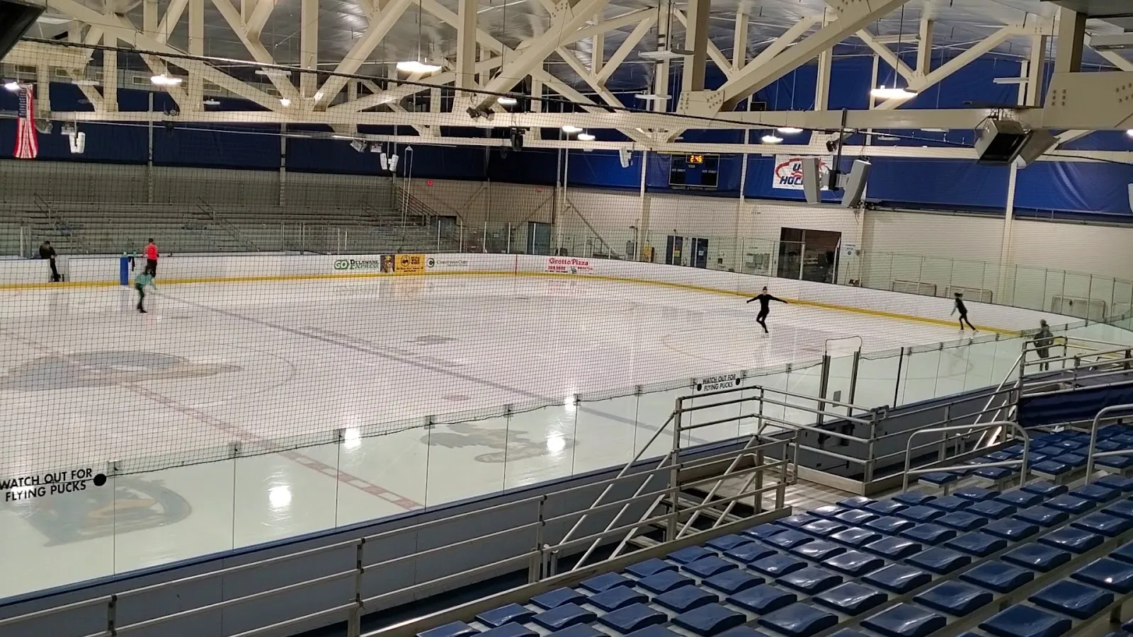 The ice at Fred Rust Ice Arena seen from the upper stands, with a few skaters far across the sheet under the white roof trusses — Fred Rust Ice Arena