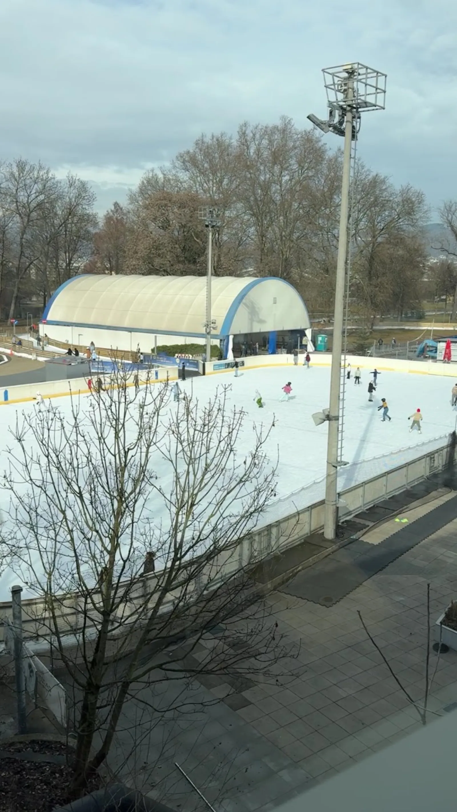 Skaters on the open-air ice at the Parkbad in Linz, with the ice hall dome behind. — Freieis und Eislaufweg Parkbad Linz