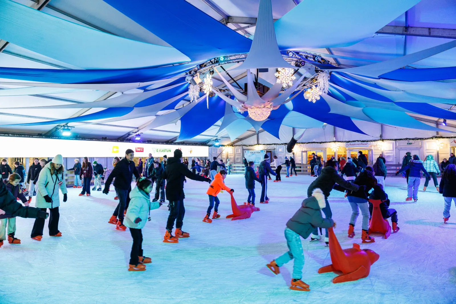 Public skating under the blue-and-white canopy of the covered Friends on Ice rink in Ede, with skaters and children using skating aids; skaters in play, not posing. — Friends on Ice