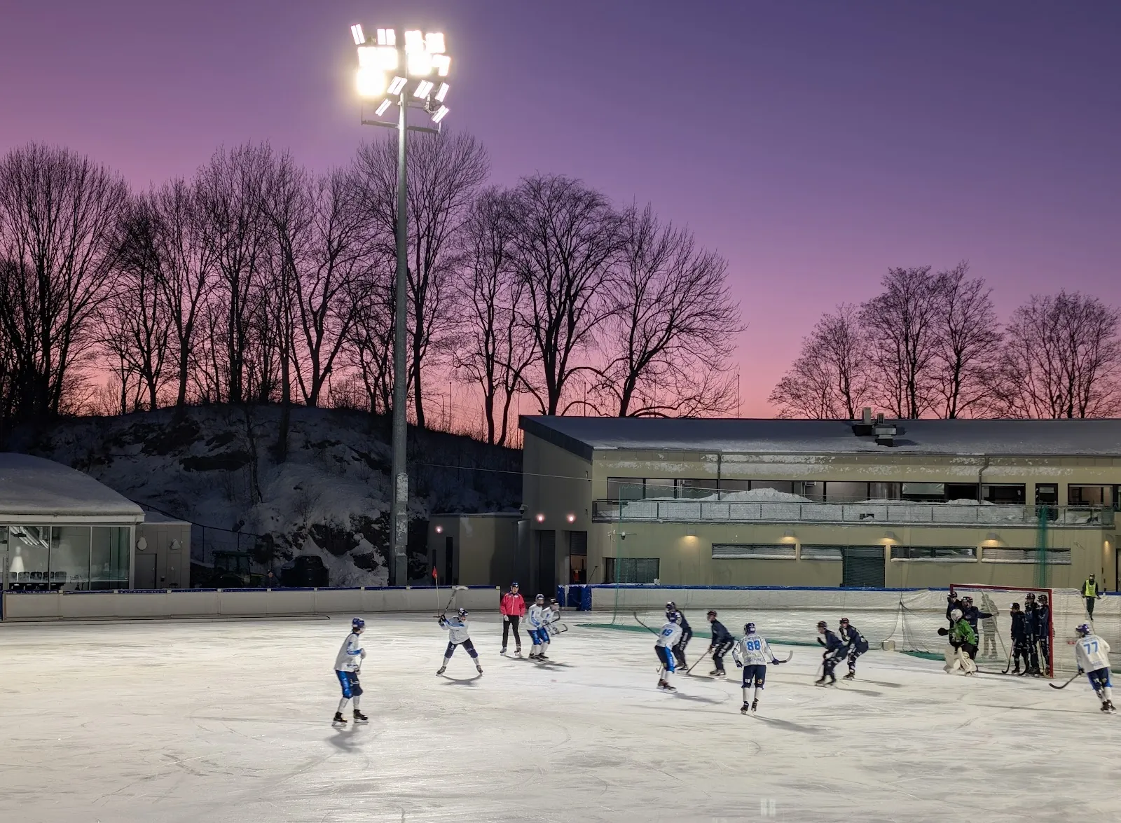 Skøyteløpere og unge hockeyspillere på utendørsisen på Frogner stadion i Oslo i skumringen, med lysmaster, tribunen og bare trær rundt banen. — Frogner stadion