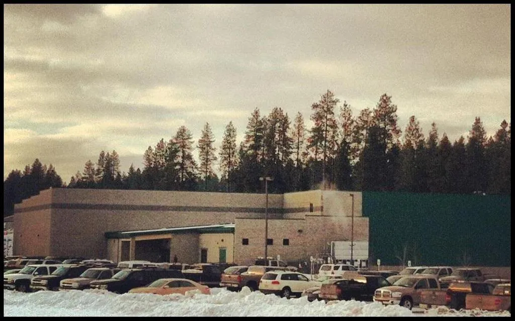The arena's tan brick building seen from the snowy parking lot, with a gabled entrance canopy, a green-paneled side wall, and evergreen trees behind it under an overcast sky. — Frontier Ice Arena