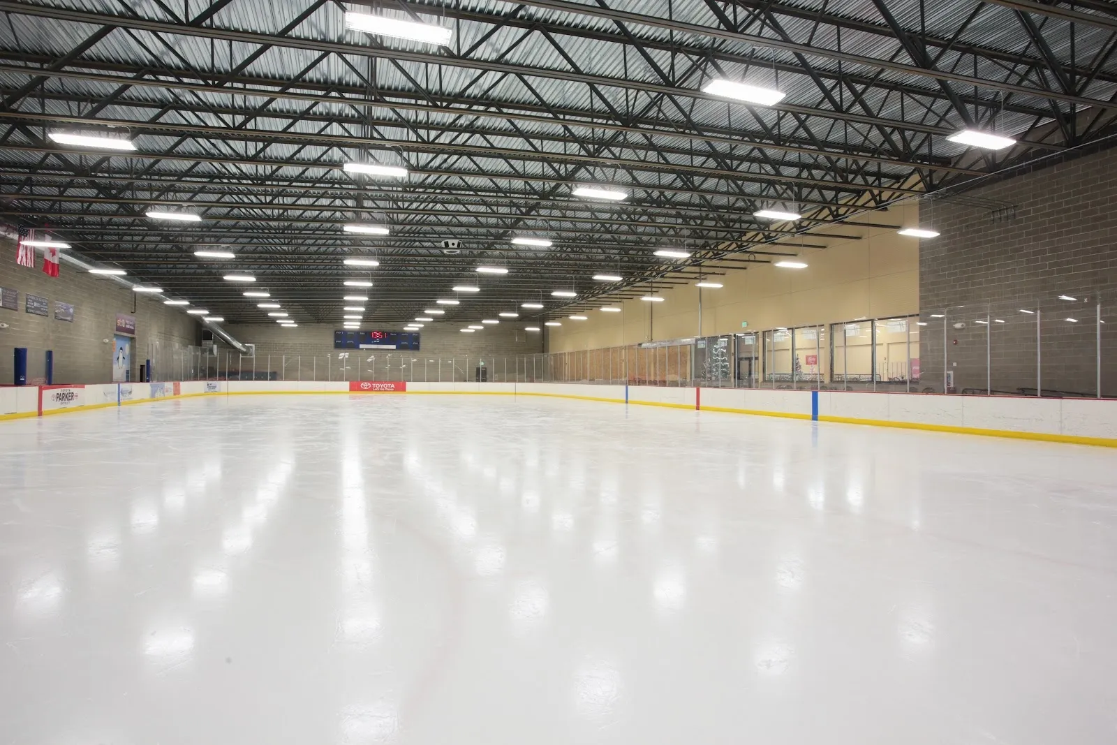 A long empty sheet seen from the end boards, white ice and yellow board line under a high roof with rows of lights and a tan wall on the right. — Frontier Ice Arena