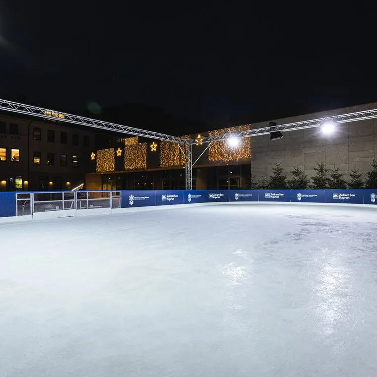 Die Freiluft-Eisbahn Frozen Daydream in Zell am See bei Nacht, mit blauer Bande unter einem Lichtgerüst und einer beleuchteten Bühne dahinter. – Frozen Daydream Eislaufplatz Zell am See