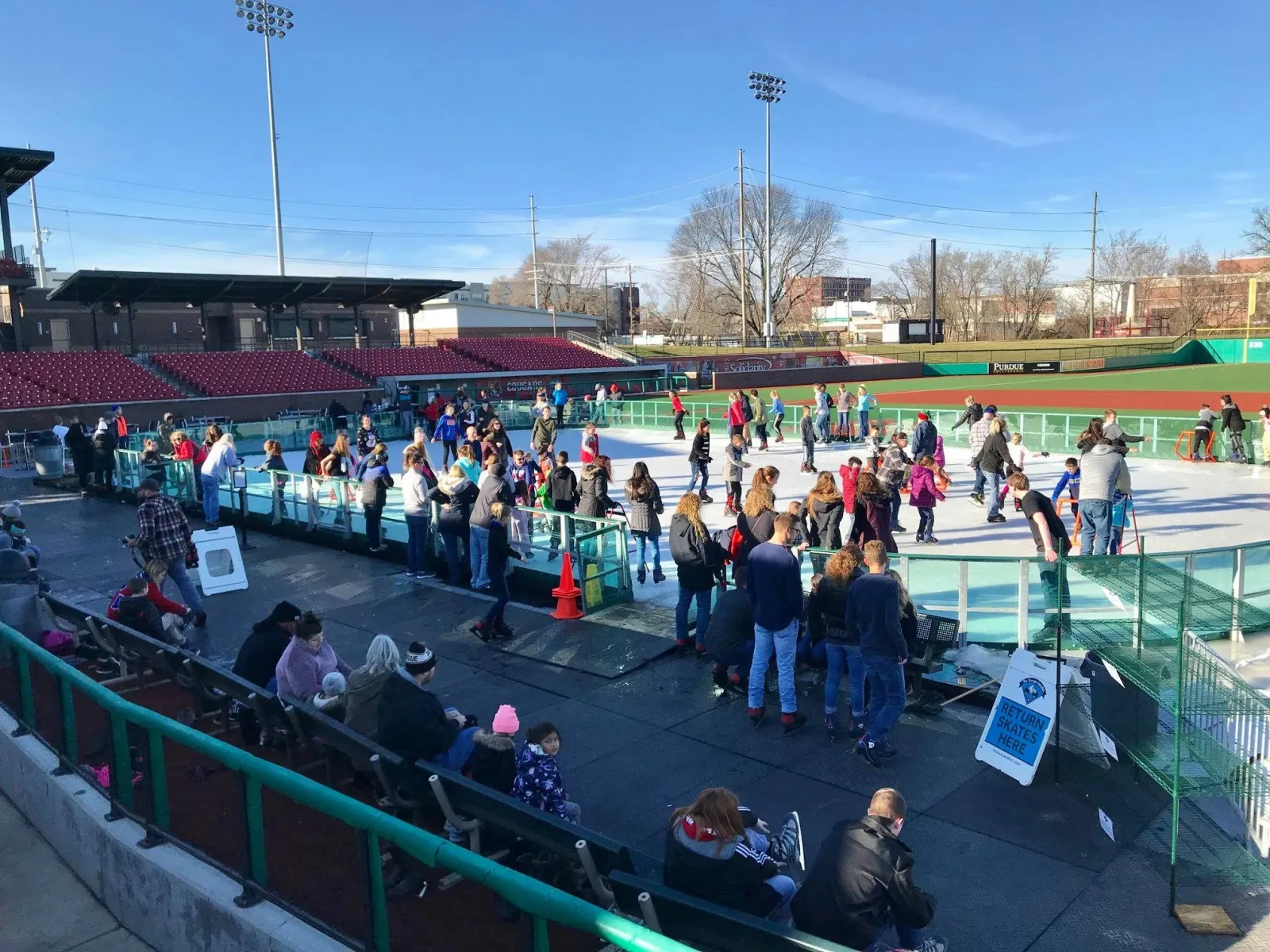 The Frozen Sandlot rink built over the ballpark infield on a sunny day, skaters and families crowding the ice with the stadium seats and light towers behind. — The Frozen Sandlot