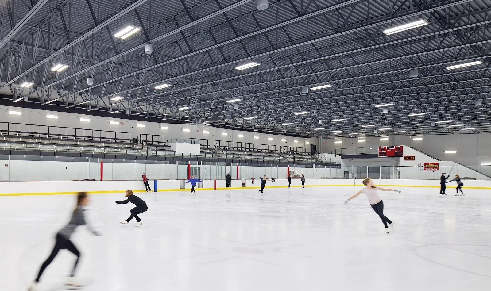 Unos cuantos patinadores repartidos por una pista blanca y amplia bajo un techo oscuro de acero, con una larga pared con frente de vidrio sobre las vallas. — Furniture and Things Community Event Center