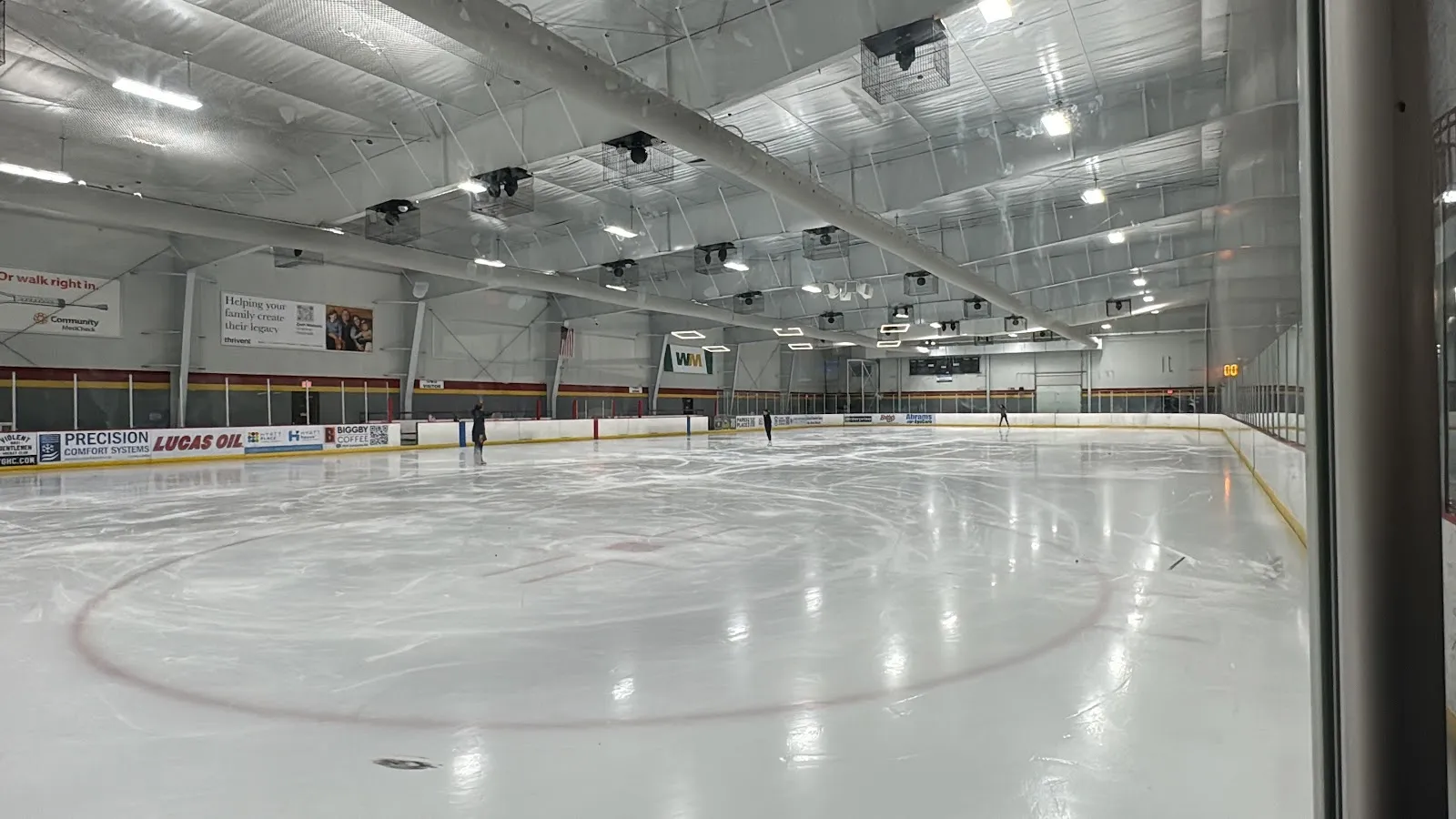A wide view of the ice through the glass at the boards, empty white sheet with centre circle under a high steel roof and benches along the near side. — Fuel Tank at Fishers