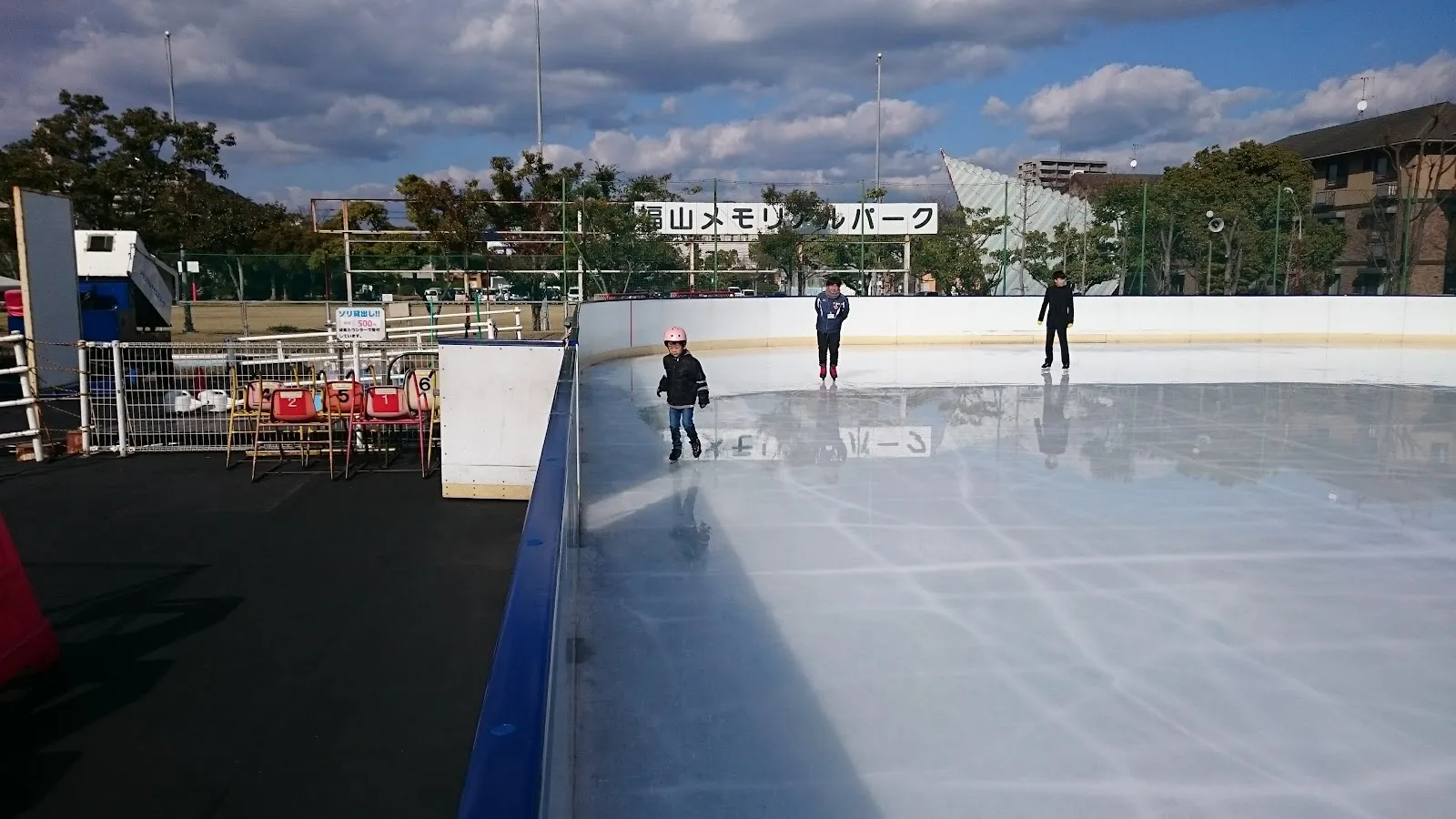 The open-air ice rink inside Fukuyama Memorial Park seen along the boards, with a few skaters at a distance, a banner reading the park's name on the fence behind, and trees and low buildings beyond. — Fukuyama Memorial Arena Ice Rink