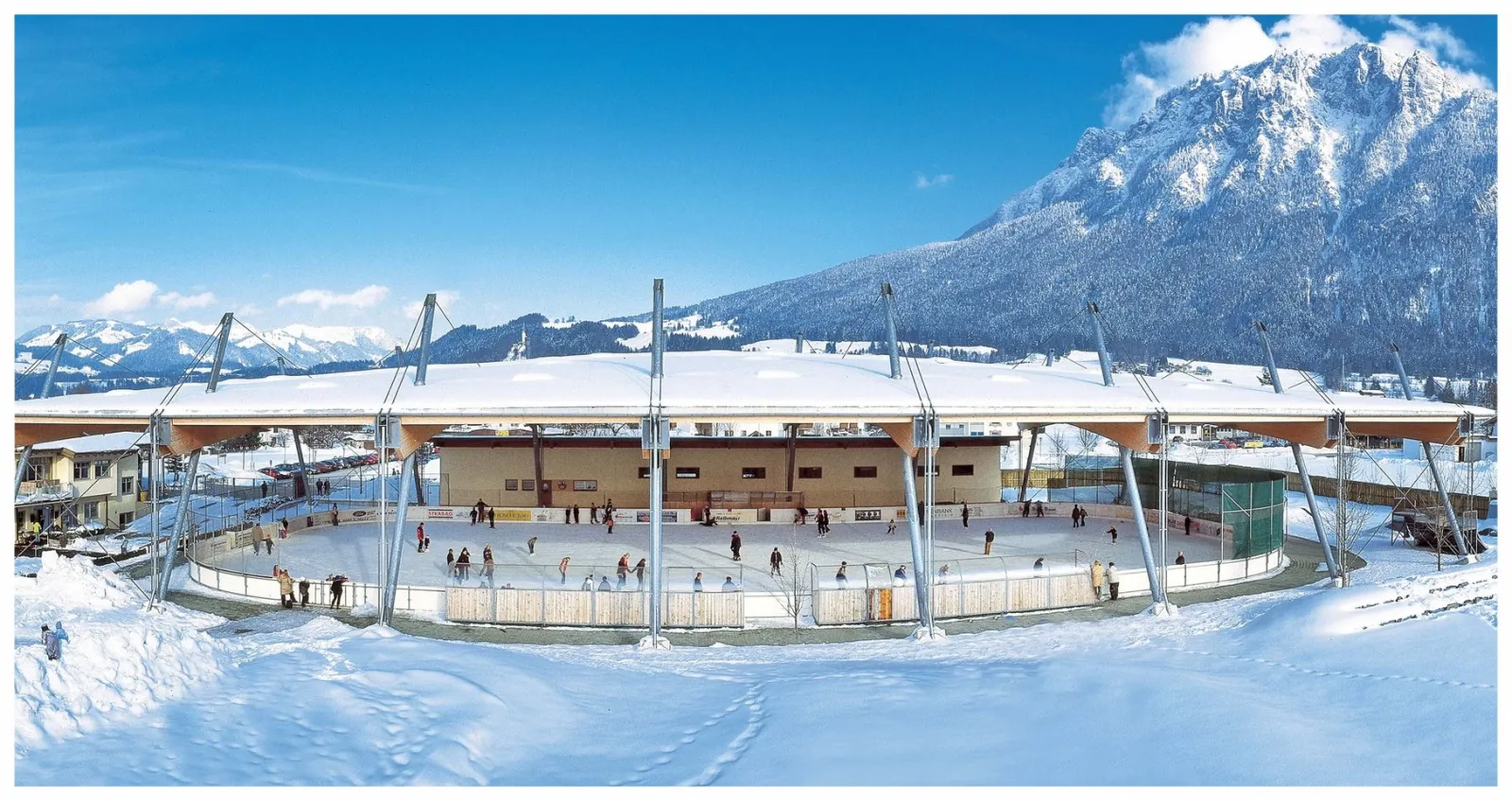 The roofed Funarena Hallo-Du in Ebbs in winter, seen from outside with snow on the roof and the Kaisergebirge mountains behind — Eislaufplatz Hallo Du Ebbs