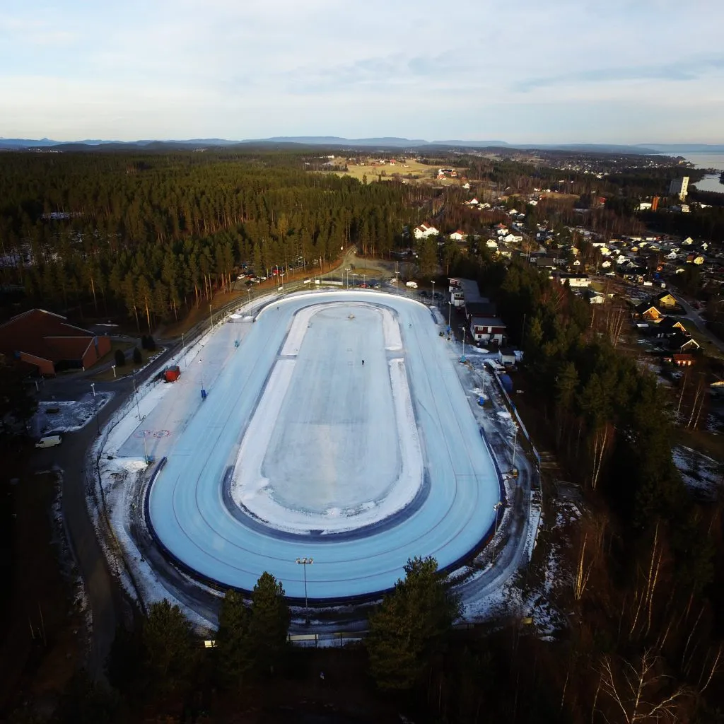 An aerial view of the Furumo speed-skating oval in winter, with the ice ring and infield surrounded by forest and the houses of Geithus. — Furumo kunstisbane