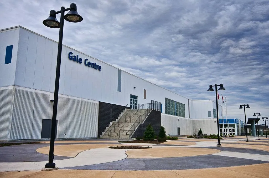 The white Gale Centre building with its blue Gale Centre lettering, an outdoor staircase, a paved plaza and lamp posts under a cloudy sky. — Gale Centre