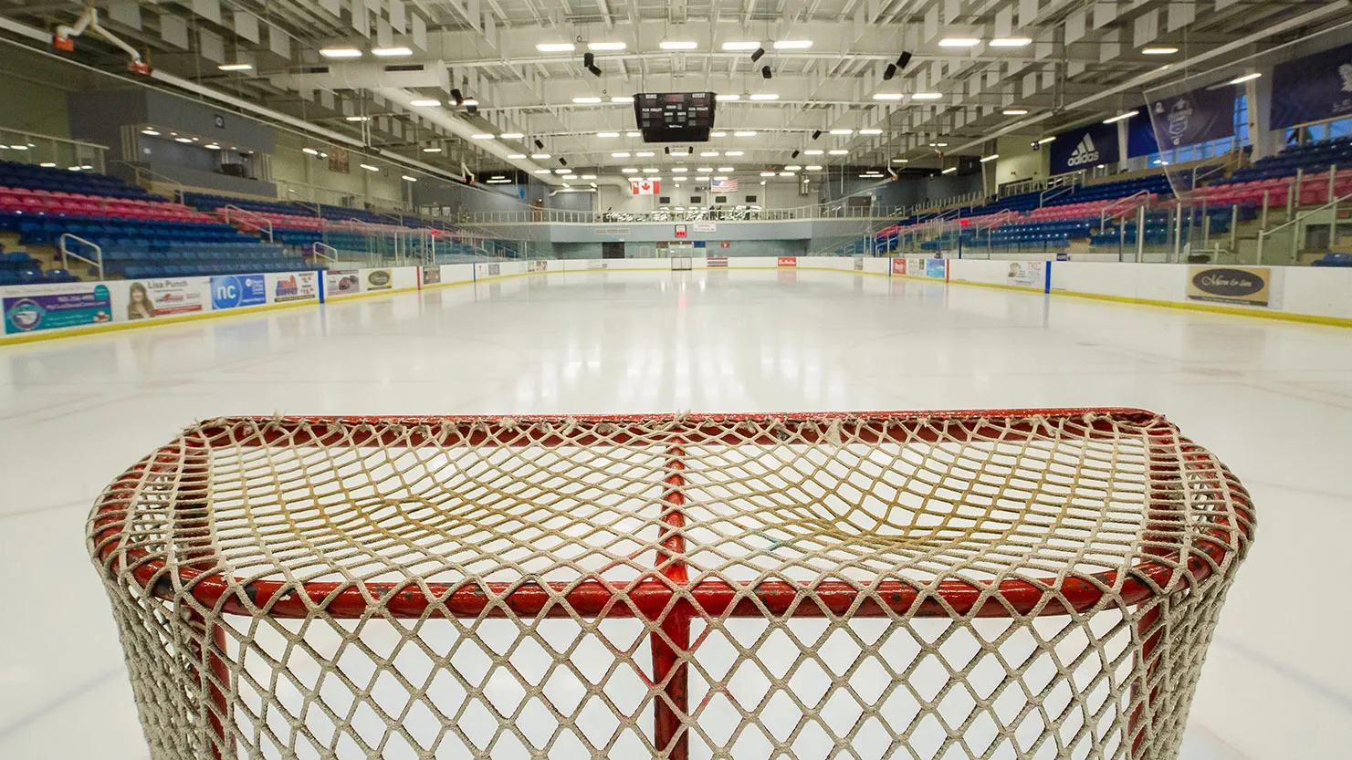 The main Gale Centre rink seen from behind a goal net, with blue seats along both sides, sponsor boards and a scoreboard hanging over the ice. — Gale Centre