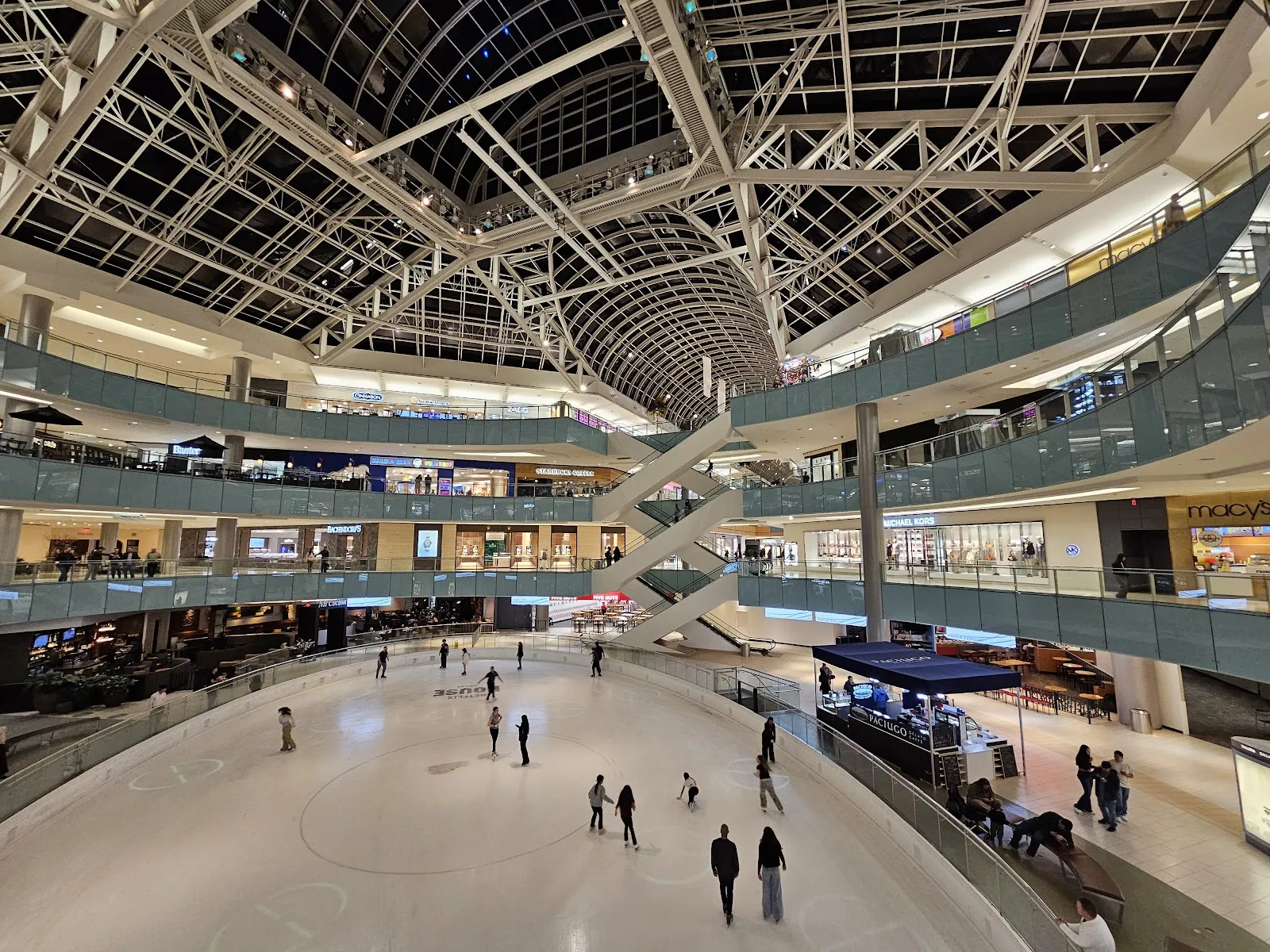 La pista vista desde el nivel superior, con patinadores repartidos por el hielo bajo el techo abovedado de vidrio y las galerías que se elevan por tres lados. — Galleria Dallas Ice Skating Center